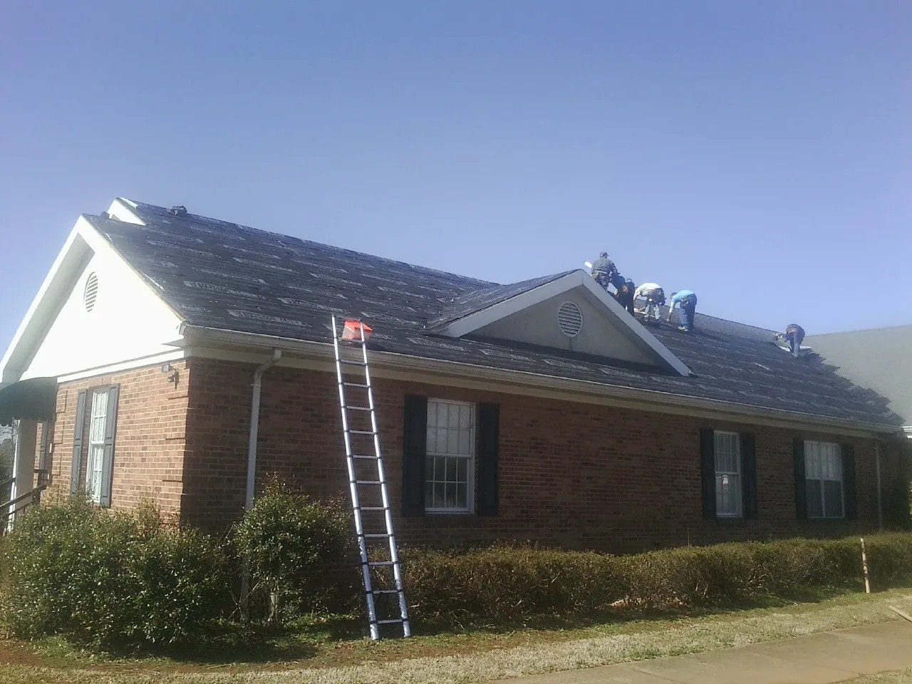 Crew tearing shingles off a brick ranch roof with a ladder set — Emergency Roof Repair work by our crew in Needham Heights