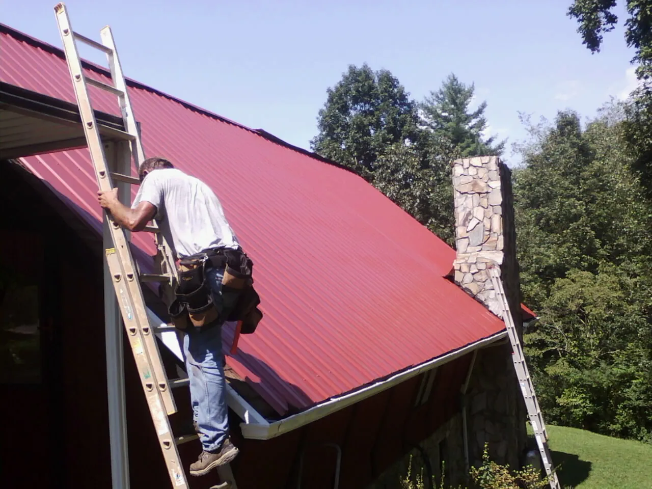 Roofer on a ladder beside a red metal roof and stone chimney — Soffit & Fascia Repair by Roofing Brothers in Needham Heights