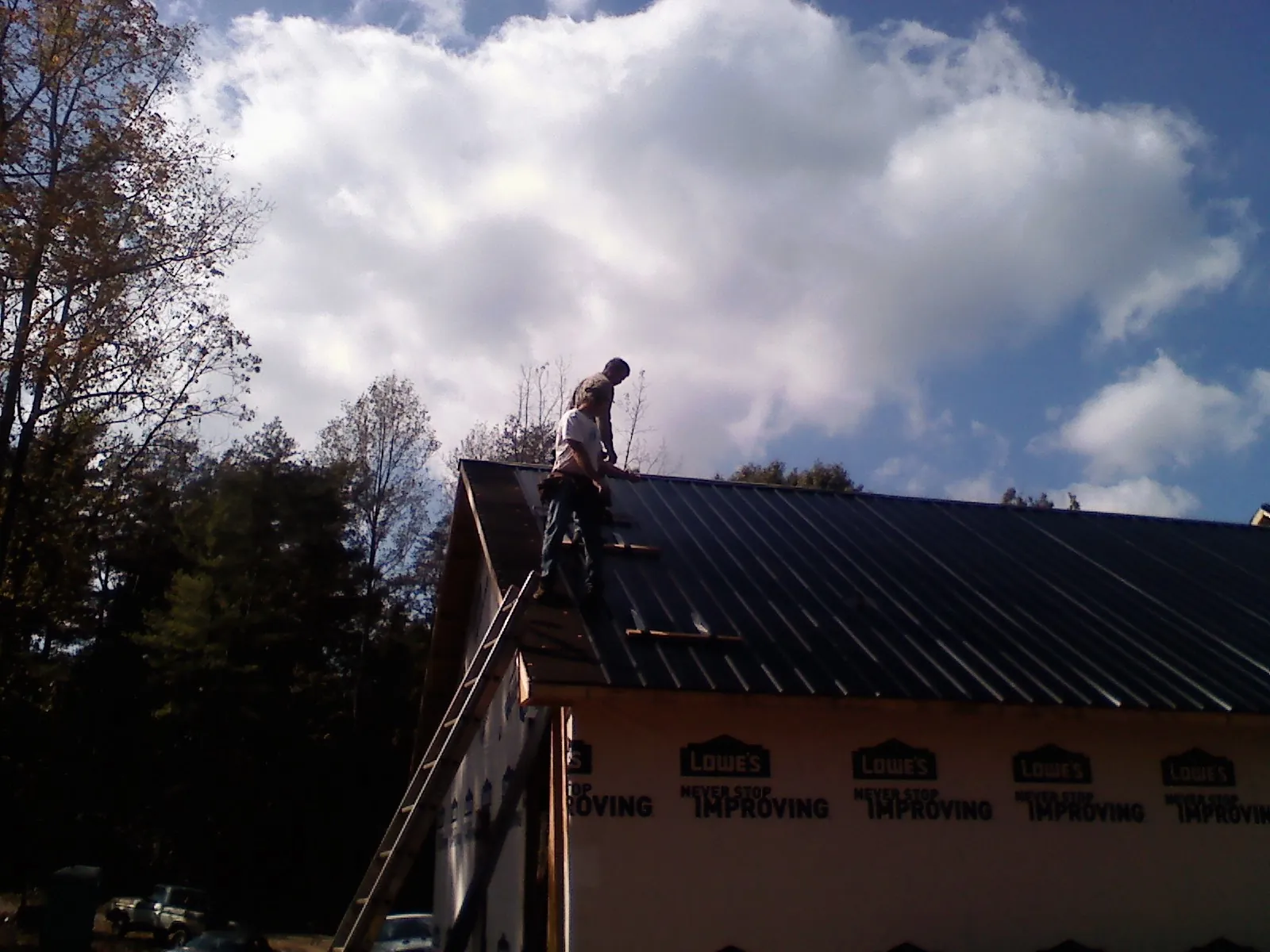 Roofer fastening dark metal panels high against a cloudy sky, from a Roof Inspection job in Needham Heights